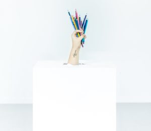 A hand is popping out of a white box holding an array of colored pencils.