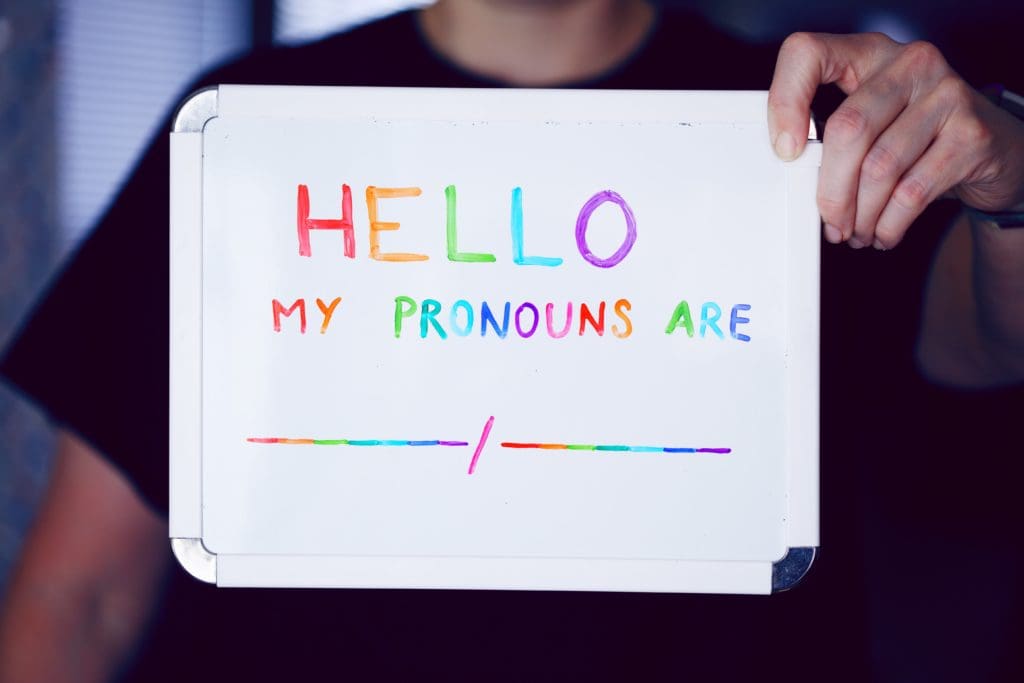 A person holding a whiteboard with colorful letters on it saying "HELLO MY PRONOUNS ARE ____/_____."