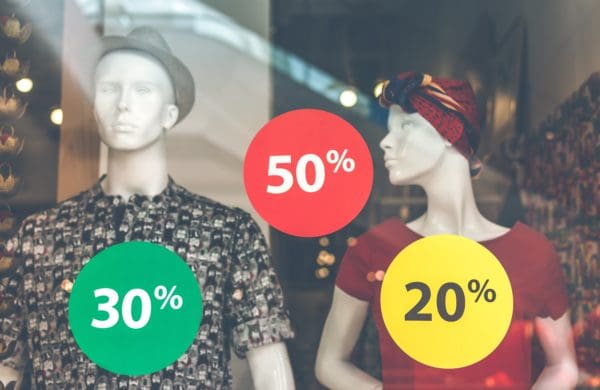 Two storefront mannequins standing beyond a glass window covered in sale stickers that say: 30%, 50%, and 20%.