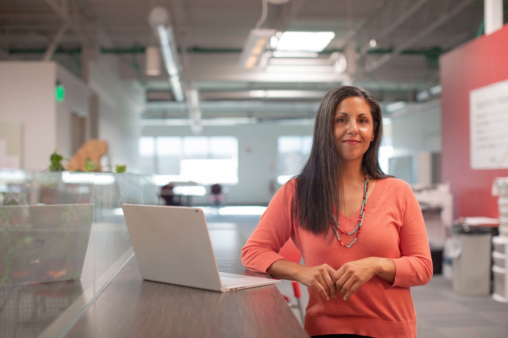 A woman stands next to a standing desk with her laptop. She is wearing a coral pink shirt.