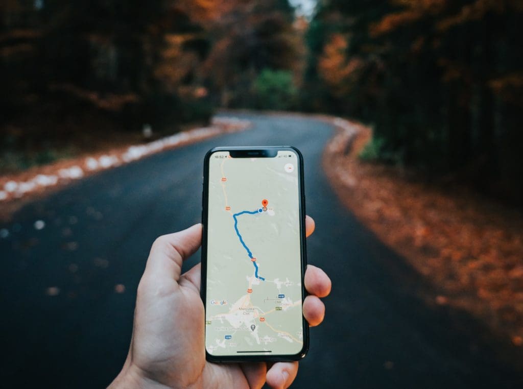 A hand holding a phone with a map application open in front a concrete path through the woods.